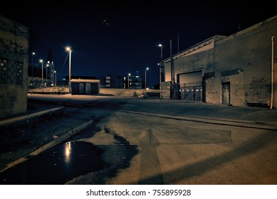 Dark Factory Warehouse Alley With Railroad Tracks And Rain Puddle At Night.