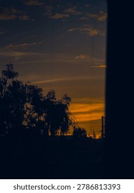 Dark, framed low angle view of tree silhouettes and utility poles against a gradient twilight sky with warm orange clouds and a dark blue horizon.