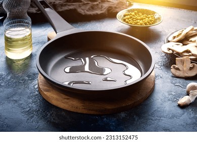 Dark frying pan with poured oil on a dark blue abstract background. Nearby is a plate of champignons, green peas, and a bottle of oil. Template for design.
