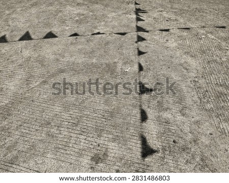 Dark, geometric shadows of festive triangular flags making a cross pattern on textured asphalt during a sunny day.