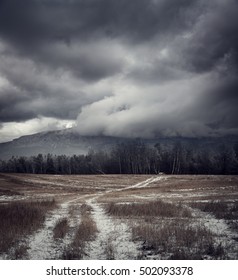 Dark Gloomy Landscape with Country Road in Snow. Moody Sky Background with Epic Dramatic Clouds around Mountains. Cold Winter Fantasy Scenery. Toned HDR Styled Photo.