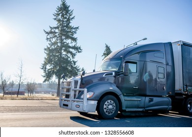 Dark Gray Big Rig American Bonnet Modern Stylish Semi Truck With Strong Grill Protection Aluminum Bumper And Covered Semi Trailer Going On The Wide Multiline Road Transporting Commercial Cargo