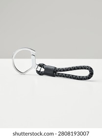 A dark grey and black patterned rope keychain with a silver metal ring rests on a white surface against a neutral light grey background, studio.