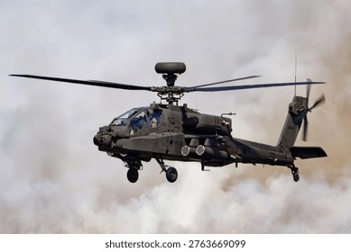 A dark grey British Army Apache AH1 helicopter, tail number ZM745, hovers with spinning rotors, enveloped by dense smoke and dust.