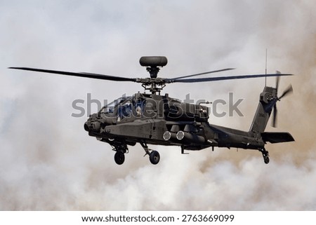A dark grey British Army Apache AH1 helicopter, tail number ZM745, hovers with spinning rotors, enveloped by dense smoke and dust.