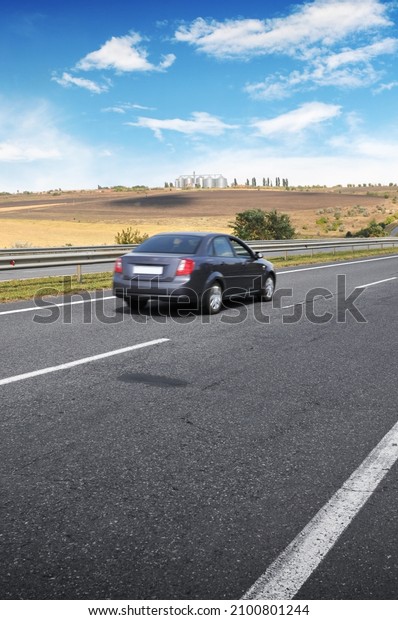 Dark grey saloon car on a countryside\
road with fields against a blue sky with\
clouds