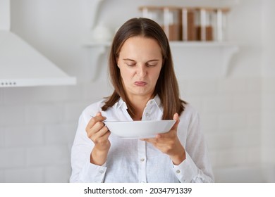 Dark Haired Caucasian Woman In White Shirt Demonstrates Disgust Twisting Face With Negative Reaction While Trying To Eat Some Smelly Soup From A Plate In Kitchen.