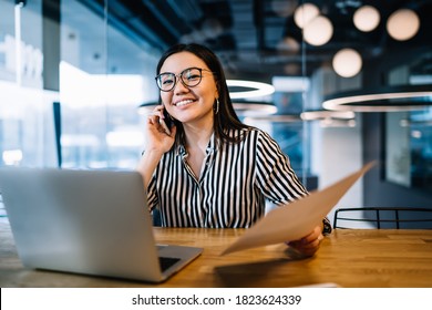 Dark Haired Cheerful Asian Woman With Glasses In Stylish Casual Clothes Sitting At Table With Laptop While Doing Paperwork And Talking On Phone