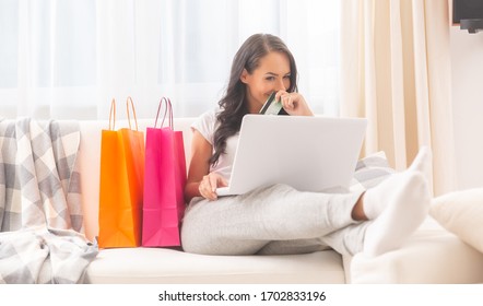 Dark haired lady dressed in comfortable light color sweatpants, and t-shirt mysteriously smiling into a laptop holding a debit card in her hand, covering mouth and with two shopping bags next to her. 