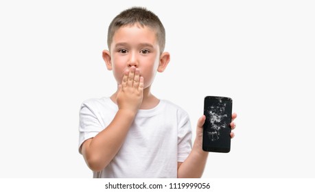 Dark haired little child holding broken smartphone cover mouth with hand shocked with shame for mistake, expression of fear, scared in silence, secret concept