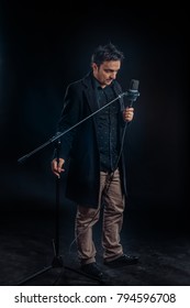 Dark haired man singing at the microphone with a dark background in studio.