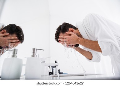 Dark Haired Man In White Bathrobe Bending His Back And Putting Both Hands On His Face While Washing It Over The Sink