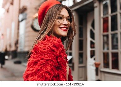 Dark Haired Tanned Woman With Red Lipstick Posing On Street. Portrait Of Young Stylish Girl In Red Knitted Coat And Hat