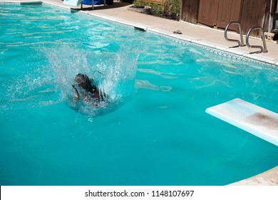 Dark Haired Woman Doing A Cannon Ball Into An Outdoor Swimming Pool In A Sunny Summer Day. Young Woman Jumps Off Diving Board Into A Backyard Swimming Pool In The Summer.