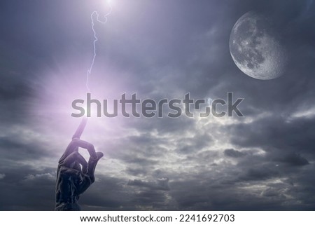 Dark hand with raised index finger in front of a heavily clouded sky with three-quarter moon from which lightning shoots. The lightning hits the index finger with bright beam.