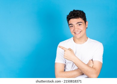 Dark hispanic teenager boy pointing to the side copy space and looking at camera isolated on blue background. Young man smiling