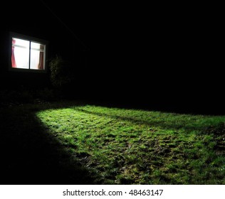 A Dark House At Night With A Window With A Light On In The Room Spilling Out Light Onto The Grass Lawn Yard