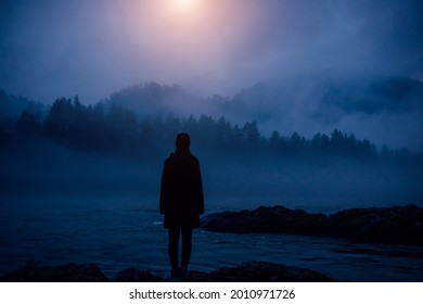 Dark human silhouette in a thick fog against the background of forest, hills and mountain river. Mysterious female figure on desert shore. Apocalyptic landscape, atmospheric pollution.