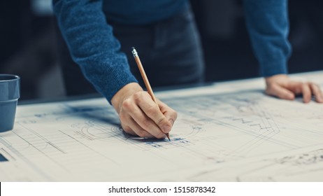 In the Dark Industrial Design Engineering Facility: Male Engineer Works and Blueprints Laying Table  Uses Pencil  Ruler   Digital Tablet  On Desktop Multiple Drawings  Focus Hands