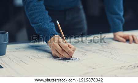 In the Dark Industrial Design Engineering Facility: Male Engineer Works with Blueprints Laying on a Table, Uses Pencil, Ruler and Digital Tablet. On Desktop Multiple Drawings. Focus on Hands