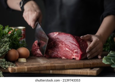 Dark and Moody. Chef in  holds a big knife and cuts into pieces raw meat on a brown wooden cutting board - Powered by Shutterstock