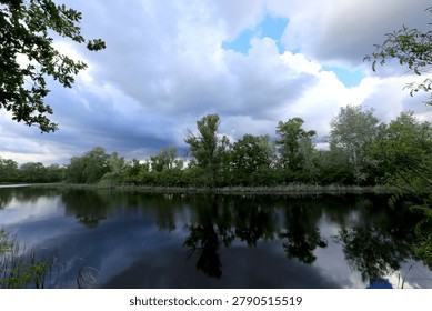 Dark, moody clouds are reflected in the still water of a lake surrounded by lush green trees and vegetation