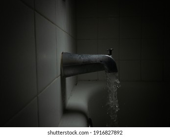 Dark, Moody Side View Of A Running Bathtub Faucet, With Water Pouring Out Into The Tub