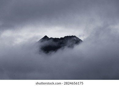 Dark mountain peak emerging through dense, moody fog and stormy gray clouds.