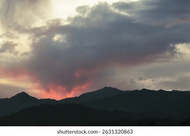 Dark mountain silhouettes beneath dramatic clouds glowing with fiery red and pink hues during a dramatic, moody sunset