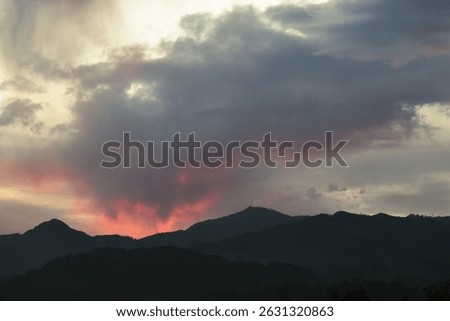 Dark mountain silhouettes beneath dramatic clouds glowing with fiery red and pink hues during a dramatic, moody sunset