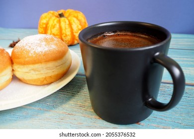 In A Dark Mug, Hot Natural Coffee Stands Next To A White Plate With A Donut And A Pumpkin On The Back Of A Blue Table. Side View