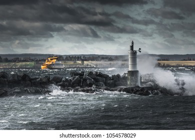 Dark and mysterious view of a yellow ferry ship struggling to hold position in stormy weather. A stone pier and a small lighthouse in the foreground with splashing waves of water