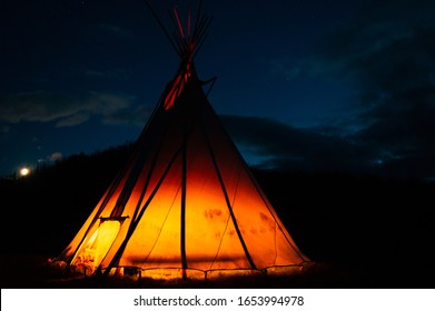 A dark night background with a tipi glowing orange from a fire inside. 