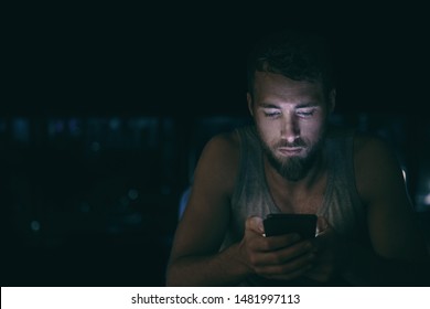 Dark Night Man Looking At Mobile Phone Screen Texting Late At Night Awake In Bed Insomnia Or Outside In City With Skyline Background. Serious Looking Guy Depressed Addicted To Social Media.
