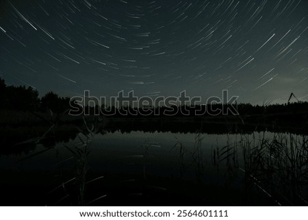 In the dark of night, star trails create a mesmerizing display above a calm lake. The still water reflects the light and surrounding trees, enhancing the tranquil atmosphere.