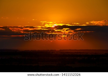 Dark orange sunset, the sun disc hides behind the clouds in the middle of the frame, casts shadows on the valley with silhouettes of fields and forests. Victory Park Ufa, Bashkortostan, Russia.