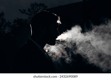 Dark photo of a silhouette of a man blowing smoke against the light. Black background and white smoke. Night portrait of a smoking man - Powered by Shutterstock