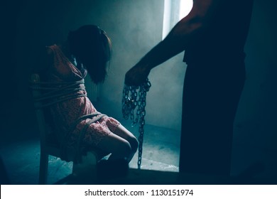 Dark picture of kidnapper standing besides his victim. He is holding chains in hand. Girl is sitting without consciousness. She is tied to chair.