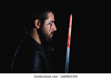 Dark Portrait Of Modern Shaman Young Man With Viking Painting. Handsome Serious Bearded Guy With Painted Face And Bloody Sword On His Hands On Black Background. Concept Of People Emotions