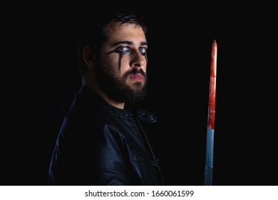 Dark Portrait Of Modern Shaman Young Man With Viking Painting. Handsome Serious Bearded Guy With Painted Face And Bloody Sword On His Hands On Black Background. Concept Of People Emotions