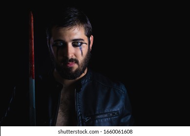 Dark Portrait Of Modern Shaman Young Man With Viking Painting. Handsome Serious Bearded Guy With Painted Face And Bloody Sword On His Hands On Black Background. Concept Of People Emotions