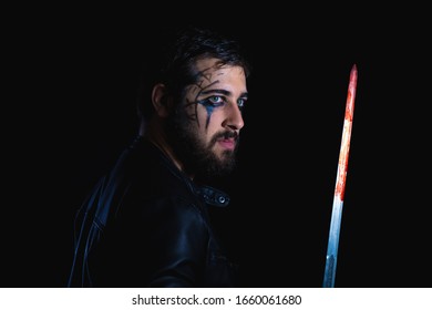 Dark Portrait Of Modern Shaman Young Man With Viking Painting. Handsome Serious Bearded Guy With Painted Face And Bloody Sword On His Hands On Black Background. Concept Of People Emotions