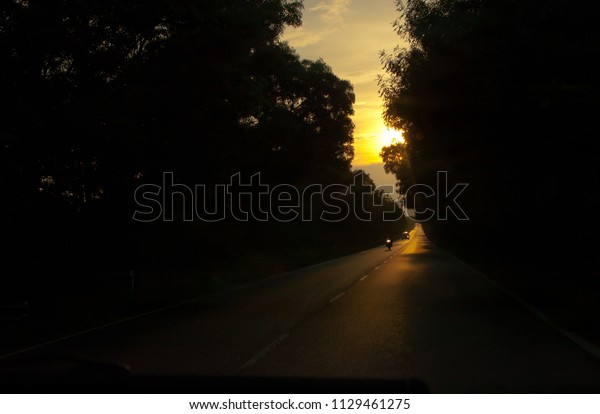 dark road at sunset through forest with\
motorcycle and car in\
distance