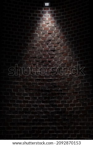 Dark room wall with ceiling light background. Old, weathered red and grey brick stone wall. Low key, scary, moody space. Interrogation room in basement. Vertical, copy space