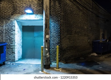 Dark And Scary Downtown Urban City Street Corner Alley With An Eerie Vintage Industrial Warehouse Factory Entrance And Dirty Dumpsters At Night