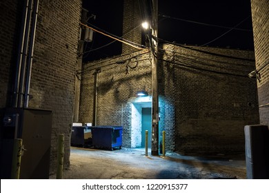 Dark And Scary Downtown Urban City Street Corner Alley With An Eerie Vintage Industrial Warehouse Factory Entrance And Dirty Dumpsters At Night