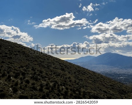 Dark scrub covered slopes frame a distant mountain and urban valley beneath sunlit cumulus clouds in Penteli, Attica, Greece
