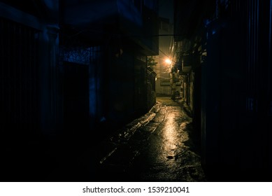 A dark, shadowy and dangerous looking urban back-alley at night time in suburbs Hanoi, Vietnam. Low light reflected on wet pavement from post lamp at the end of long road corner - Shutterstock ID 1539210041