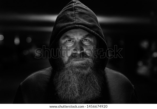 Dark shadowy portrait of a bearded man wearing\
a hooded top staring intently at the camera with a focused\
determined expression