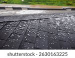 Dark shingled roof covered with scattered small hailstones during a hailstorm, with a green lawn in the background. High quality photo